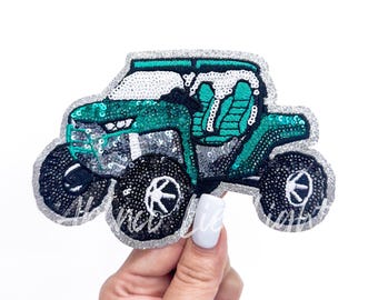 May include: A sequined patch in the shape of a teal and silver off-road vehicle. The patch has black wheels with white accents and a silver glitter border. The vehicle's interior is teal, and the roof is white.