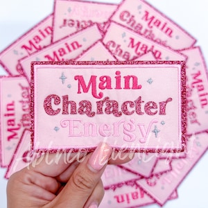 May include: Pink and red glitter iron-on patch with the text "Main Character Energy" in a pink and red font.