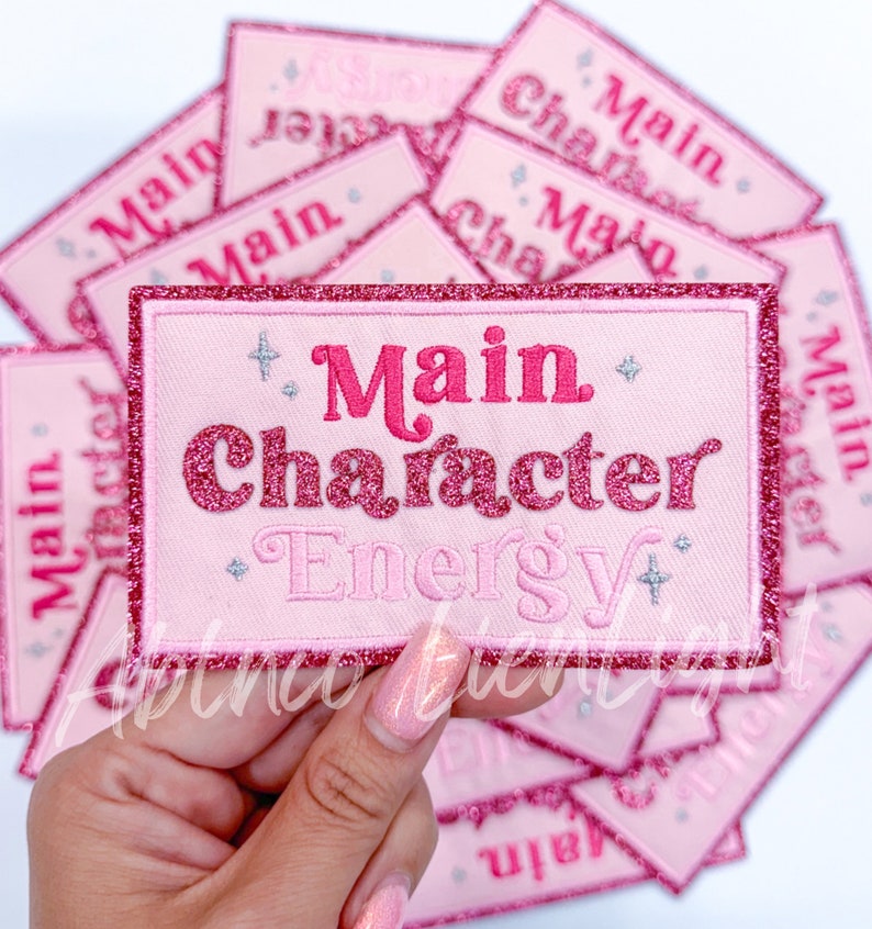 May include: Pink and red glitter iron-on patch with the text "Main Character Energy" in a pink and red font.