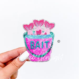 May include: A pink sequined bucket with the word "BAIT" in turquoise sequins. The bucket contains three pink mermaid tails with heart-shaped fins. The design is on a white background.