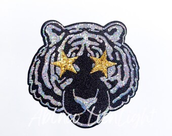 May include: A black sequined tiger face with gold star-shaped eyes. The tiger's stripes are silver sequins. The image is on a white background. The text "Abino Moon Light" is at the bottom.