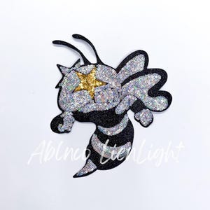 May include: A black and silver sequined bee with a gold star on its head. The bee is in a cartoon style and has a friendly expression.