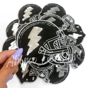 May include: A close-up of a stack of black and silver sequined iron-on patches shaped like football helmets with a lightning bolt design.