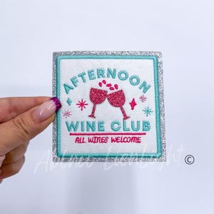 May include: A white iron-on patch with a silver glitter border. The patch features a turquoise blue background with the text "Afternoon Wine Club" in white. The text "All wines welcome" is in smaller white text below the main text. Two pink wine glasses with pink glitter are clinking together with pink glitter falling from the glasses.