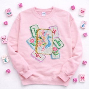 May include: A light pink sweatshirt with a colorful embroidered design of Mahjong tiles. The central tile features a blue bird and floral accents. Pink dice and decorative tiles surround the sweatshirt.