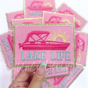 May include: A stack of pink embroidered iron-on patches with gold glitter trim. The patches feature a pink boat with a yellow sun and the text "LAKE LIFE" in turquoise.