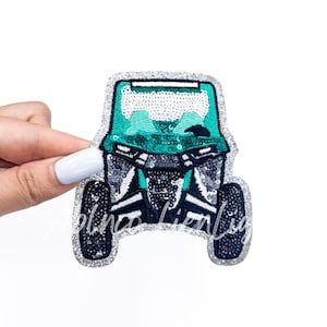 May include: A sequined patch in the shape of a teal and black off-road vehicle. The patch has a silver glitter border and is on a white background. The vehicle has a roll cage and large tires.