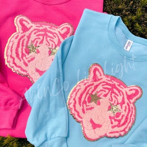 May include: Two sweatshirts with a pink glitter tiger head design. One sweatshirt is pink and the other is light blue.