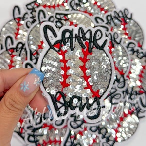 May include: A stack of white and black baseball-themed iron-on patches with silver sequins. The top patch features the word "Game" in black cursive lettering and the word "Day" in black block lettering. The patches are perfect for adding a sporty touch to clothing, bags, or other accessories.