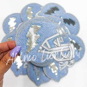 May include: A stack of blue felt patches with a silver sequin lightning bolt and a white embroidered football helmet.