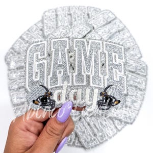 May include: Silver glitter iron-on patch with the text "GAME day" and two football helmets.