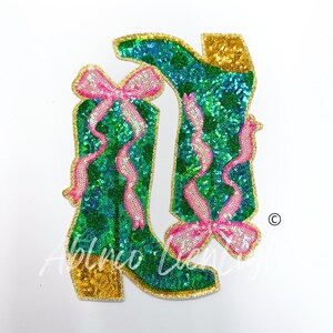 May include: A pair of green sequined boots with pink bows. The boots have gold trim and are decorated with pink ribbons.