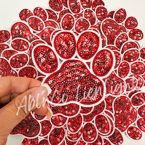 May include: A pile of red and white paw print iron-on patches with sequins. The patches are arranged in a fan shape.