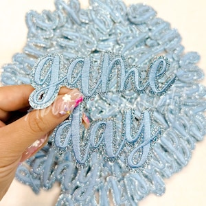 May include: Light blue embroidered patches with the words "Game Day" in a cursive font. The patches have a glittery silver border and are scattered on a light blue background.