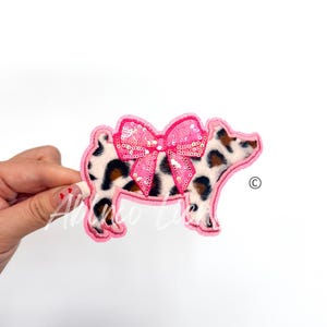May include: Pink and brown pig patch with a pink sequined bow. The pig is wearing a leopard print fur pattern.