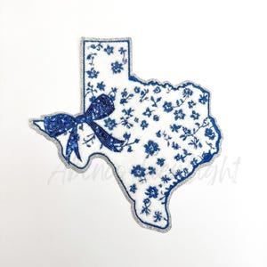May include: A white and blue embroidered patch in the shape of the state of Texas with a blue bow. The patch has a silver glitter outline.