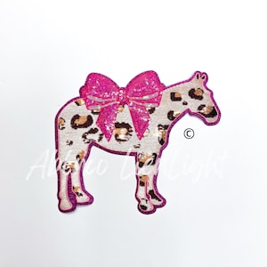 May include: A decorative patch in the shape of a horse with a pink sequined bow. The horse has a white background with brown leopard spots. The patch is outlined in pink.