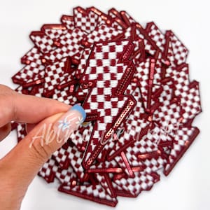 May include: A stack of red and white checkered lightning bolt patches with pink sequins. The patches are made of fabric and are about 2 inches tall.
