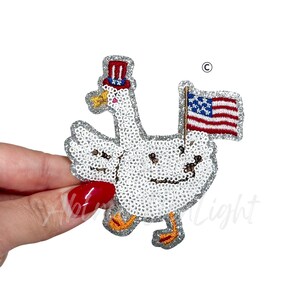 May include: A white sequined goose patch with a patriotic theme. The goose wears a red, white, and blue top hat and carries a small American flag. The patch has silver glitter edging and orange feet.