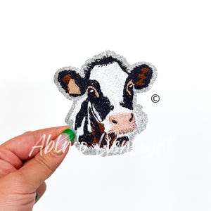 May include: A black and white cow with brown ears, a pink nose, and a white background. The cow is embroidered with glitter.