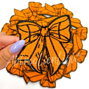 May include: An orange bow-shaped patch with black glitter detailing. The patch is held in front of a collection of similar orange bow patches. The bow has a detailed embroidered design.