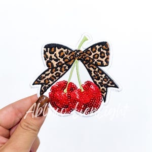 May include: Embroidered patch featuring two red sequined cherries with green stems, topped with a leopard print bow. The patch has a white border and is on a white background.