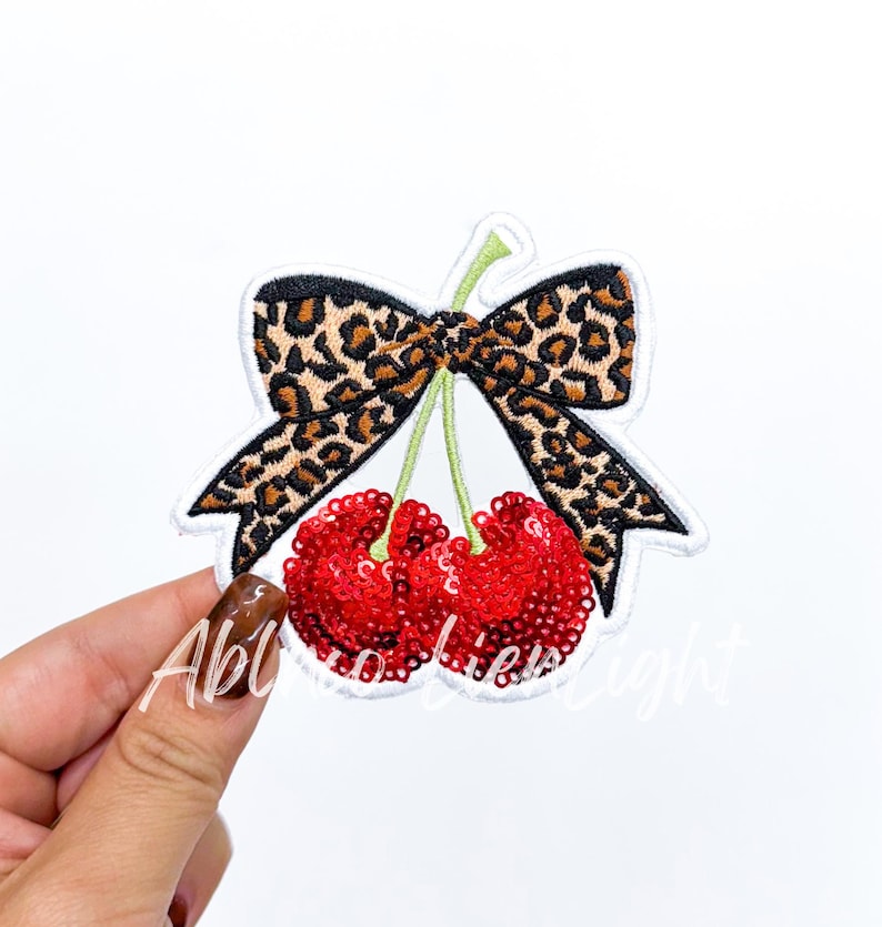 Puede incluir: Parche bordado con dos cerezas rojas con lentejuelas y tallos verdes, coronadas con un lazo con estampado de leopardo. El parche tiene un borde blanco sobre un fondo blanco.