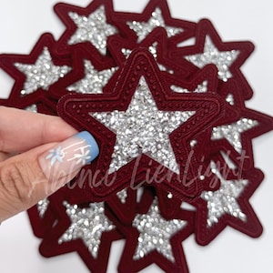 May include: A pile of burgundy red felt star-shaped patches with a silver glitter star design in the center.