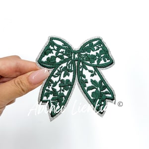 May include: A green embroidered bow with a silver glitter outline. The bow is a delicate design with floral details.
