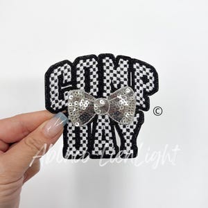 May include: Black and white checkered fabric patch with the text "COMP DAY" and a silver sequin bow.