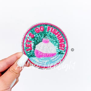 May include: A round, sequined patch with the words "LET'S GO FISHING!" in pink lettering. The patch features a white and pink bobber design on a teal background, with a silver border.