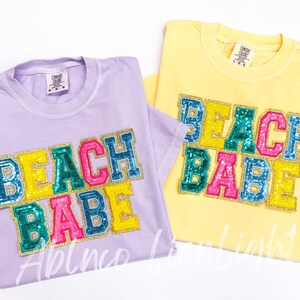May include: Two t-shirts with the words "Beach Babe" in colorful, glittery letters. The shirts are in lavender and yellow.