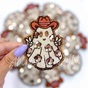 May include: A white ghost wearing a brown cowboy hat with a red band, decorated with gold sequins. The ghost is holding up two fingers in a peace sign on each hand.