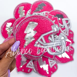 May include: A close-up of a pink and silver sequined football helmet patch. The helmet is outlined in white and has a silver lightning bolt on the side.