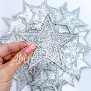 May include: Silver glitter star-shaped patches. The patches have a silver glitter surface and a silver border. A hand is holding one of the patches. Other patches are in the background.