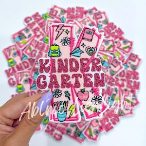 May include: A pink and white iron-on patch with the text "KINDERGARTEN" in pink glitter letters. The patch is decorated with various school-themed illustrations, including a backpack, pencils, a lightning bolt, and a heart.
