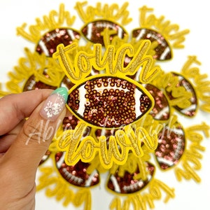 May include: A close-up of a gold and brown iron-on patch with the words "touch down" and a sequined football. The patch is surrounded by other similar patches.