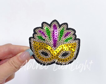 May include: A gold and purple sequined Mardi Gras mask with green and purple feathers. The mask is on a white background.