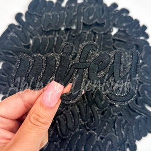 May include: Black iron-on patch with silver glitter outlining the word "wifey".