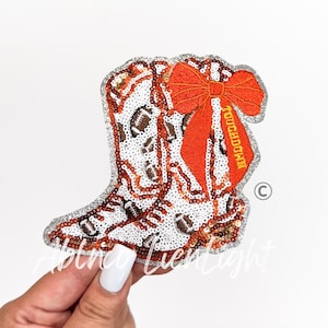 May include: A sequined iron-on patch shaped like a cowboy boot, featuring a white background with brown football patterns, a large orange fabric bow, and the word TOUCHDOWN embroidered in yellow thread on the side, finished with silver trim.
