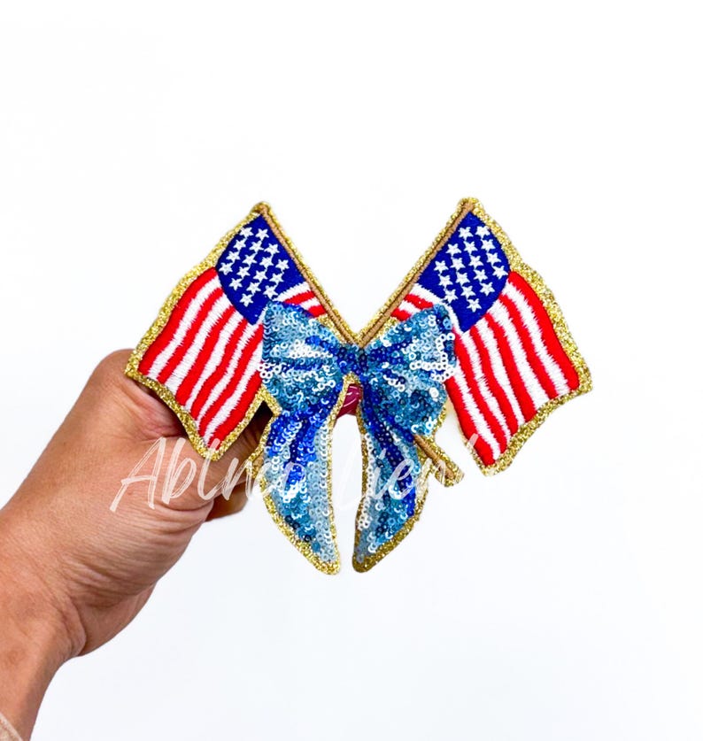 May include: Two American flags crossed over each other with a blue and gold sequined bow in the center. The flags are red, white, and blue with gold trim.