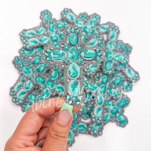 May include: A turquoise and silver embroidered cross with a beaded design. The cross is held in a hand with light green nail polish.