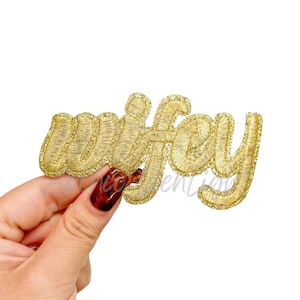 May include: A gold embroidered patch with the word "wifey" in a cursive script. The patch has a glittery texture and is held against a white background.