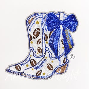 May include: White cowboy boot with brown footballs and gold stars, embellished with sequins. A large blue sequined bow is attached to the boot. The boot has a brown heel and a silver glitter trim. The design is on a white background.