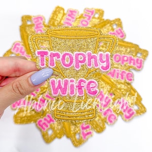 May include: A gold glitter trophy with the words "Trophy Wife" in pink glitter lettering. The trophy is surrounded by other similar trophies.