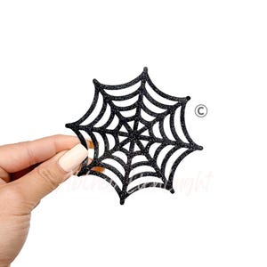 May include: A black glitter spiderweb decoration. The spiderweb is held against a white background. The spiderweb is a Halloween-themed decoration.