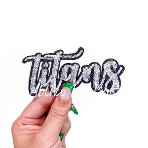 May include: A sequined patch in the shape of the word "Titans" in a cursive font. The letters are filled with silver sequins and outlined in black glitter. The patch is held against a white background.