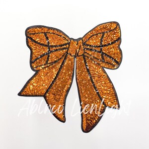 May include: A large orange sequined bow with a black outline. The bow is made of sequins and has a shiny finish.