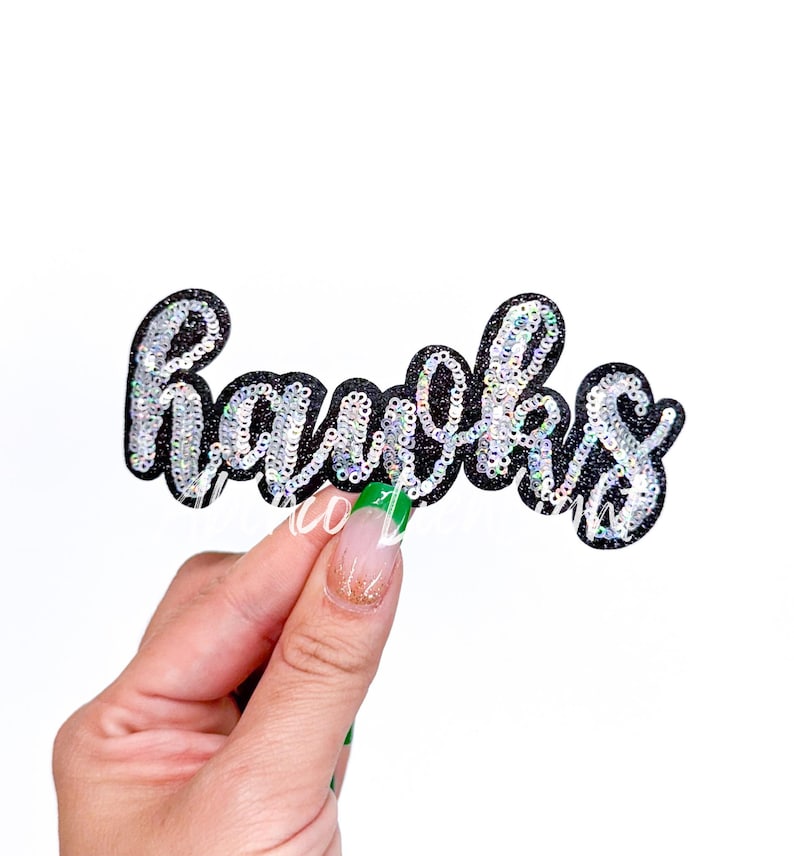 May include: A black glittery patch with the word "Hawks" in silver sequins. The patch is held against a white background. The patch is a decorative item.
