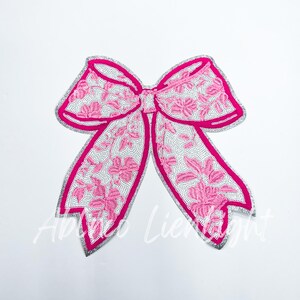 May include: A pink and white bow with floral embroidery and silver trim. The bow is made of fabric and has a shiny finish.